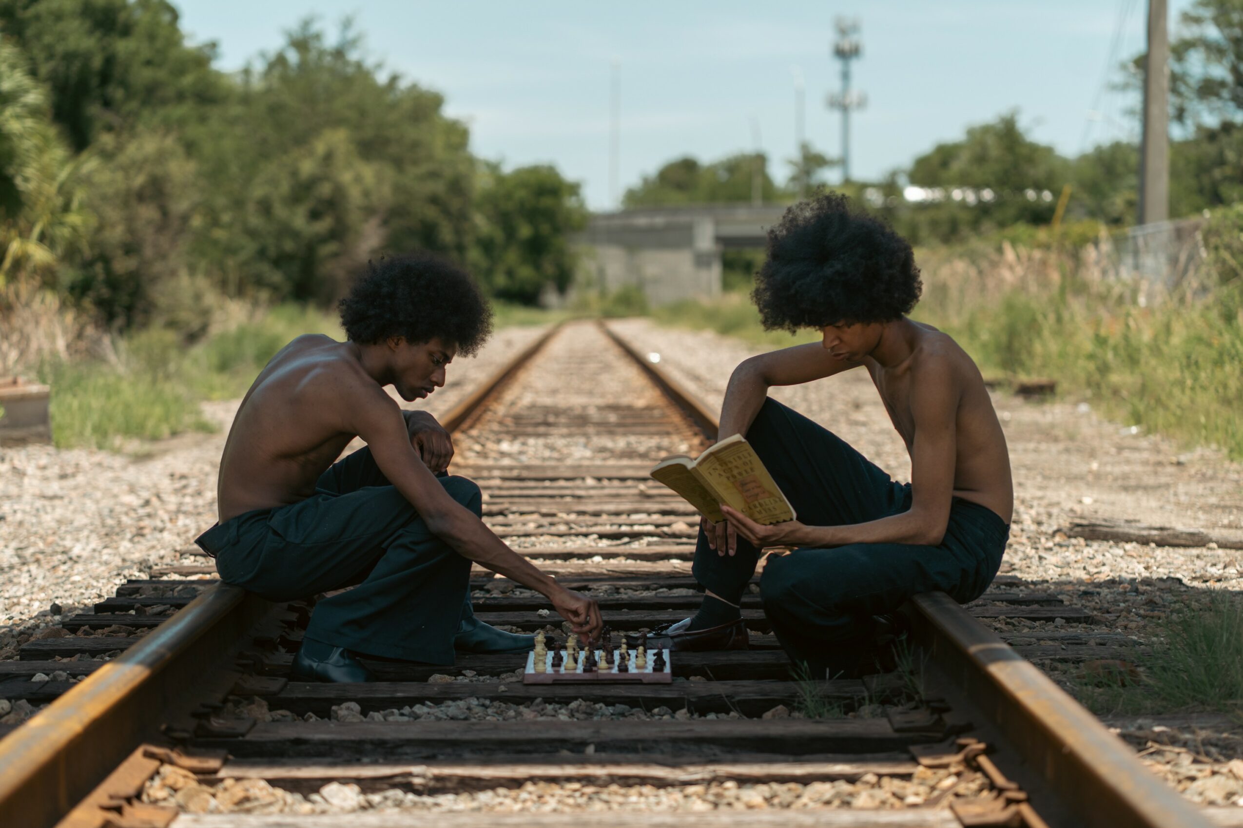 Two children with curly hair sit on train tracks, one reading a book, the other playing chess, outdoors on a sunny day.
