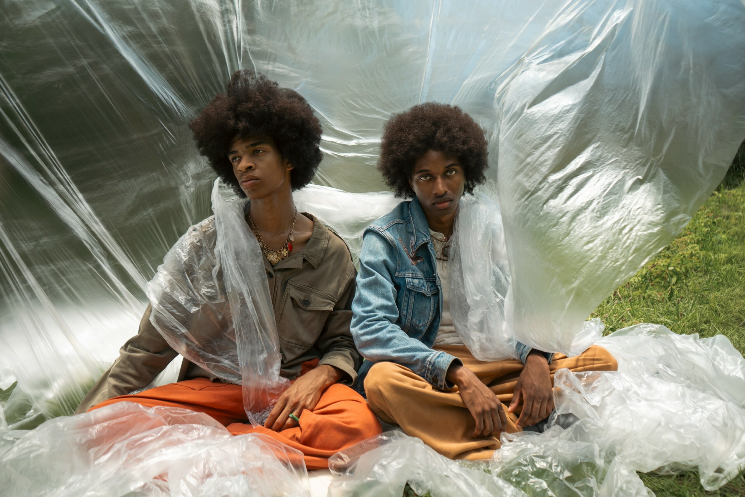 Two people with curly hair sit on the ground, surrounded by transparent plastic sheets, outdoors with greenery in the background.