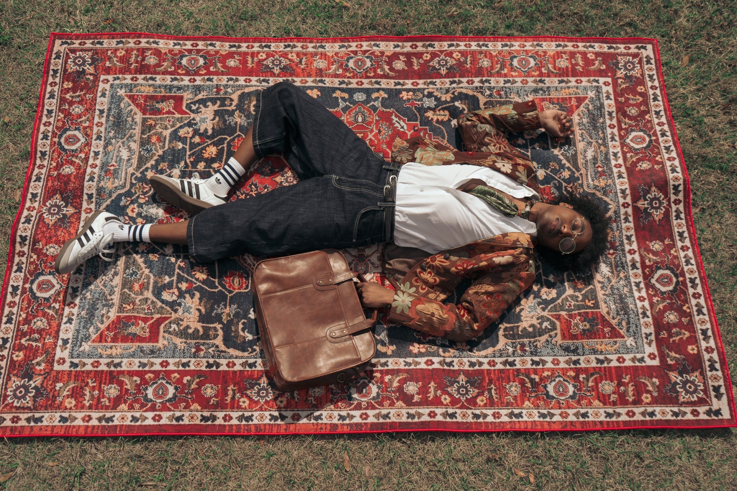 Person lying on a patterned rug outdoors, wearing a jacket, white shirt, black pants, and sneakers, with a bag nearby.