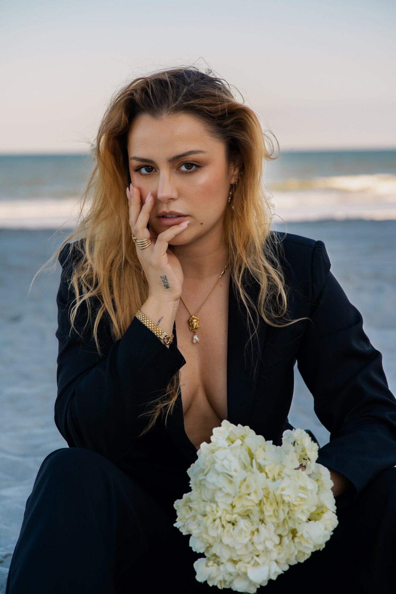 Woman with wavy hair in a black outfit holding a white flower bouquet at the beach, with ocean and sky in background.