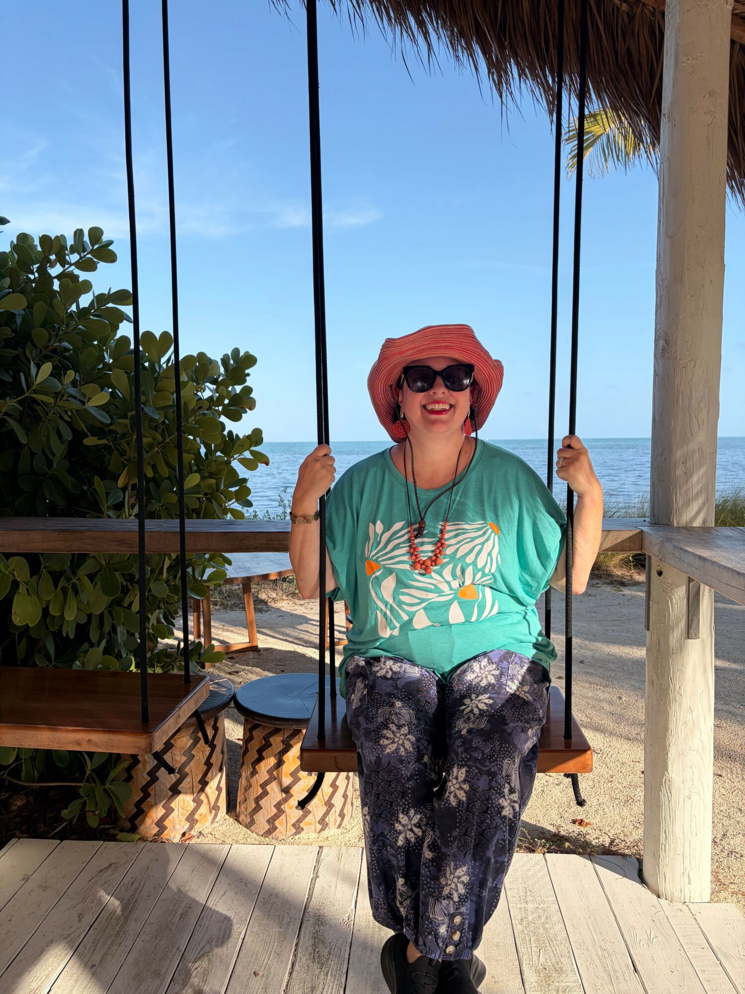 Person wearing sunglasses, a pink hat, turquoise shirt, and patterned pants, standing on a wooden deck with a swing and greenery.