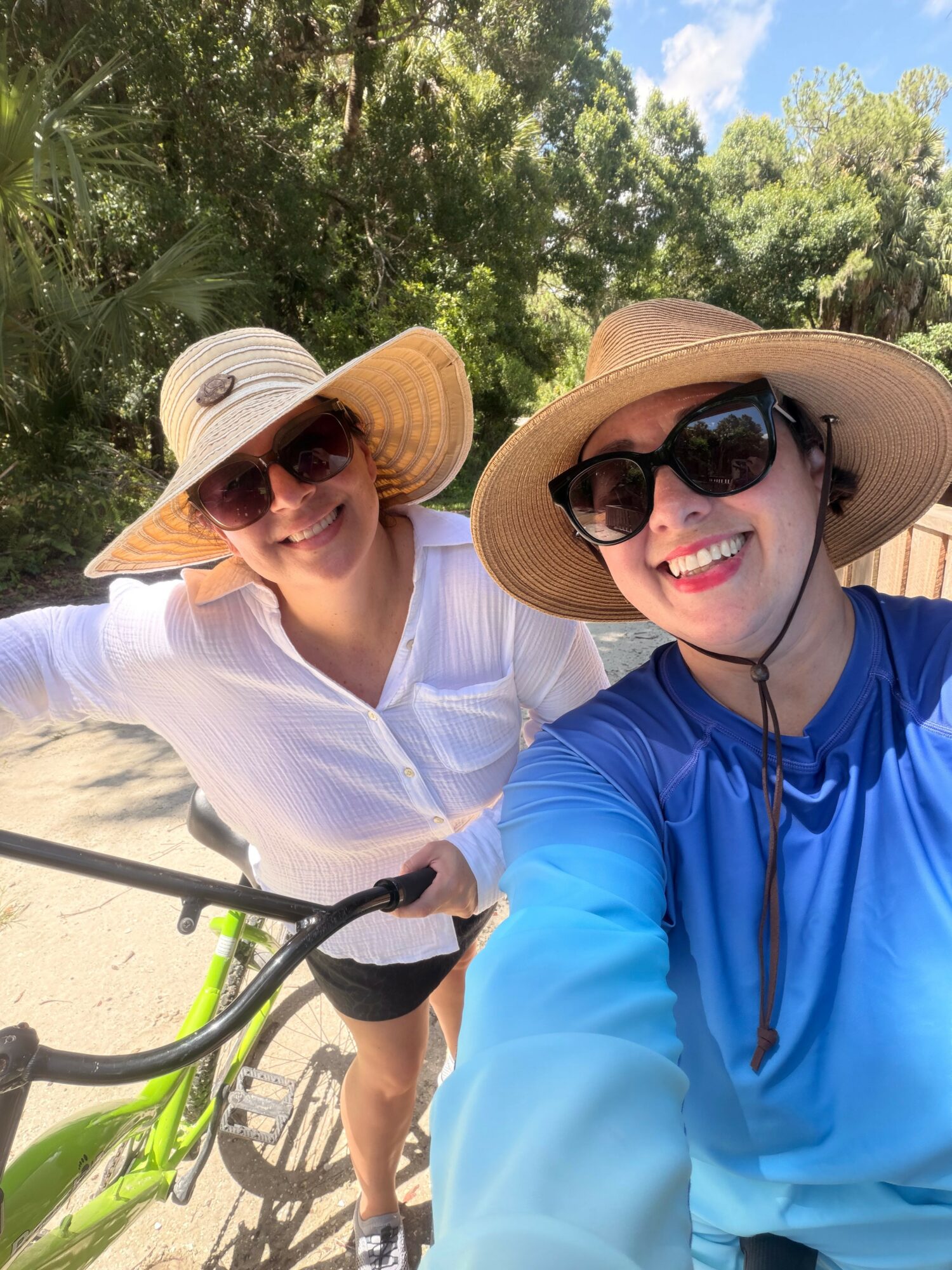 Two women outdoors wearing wide-brimmed hats and sunglasses, smiling, with trees and blue sky in background.