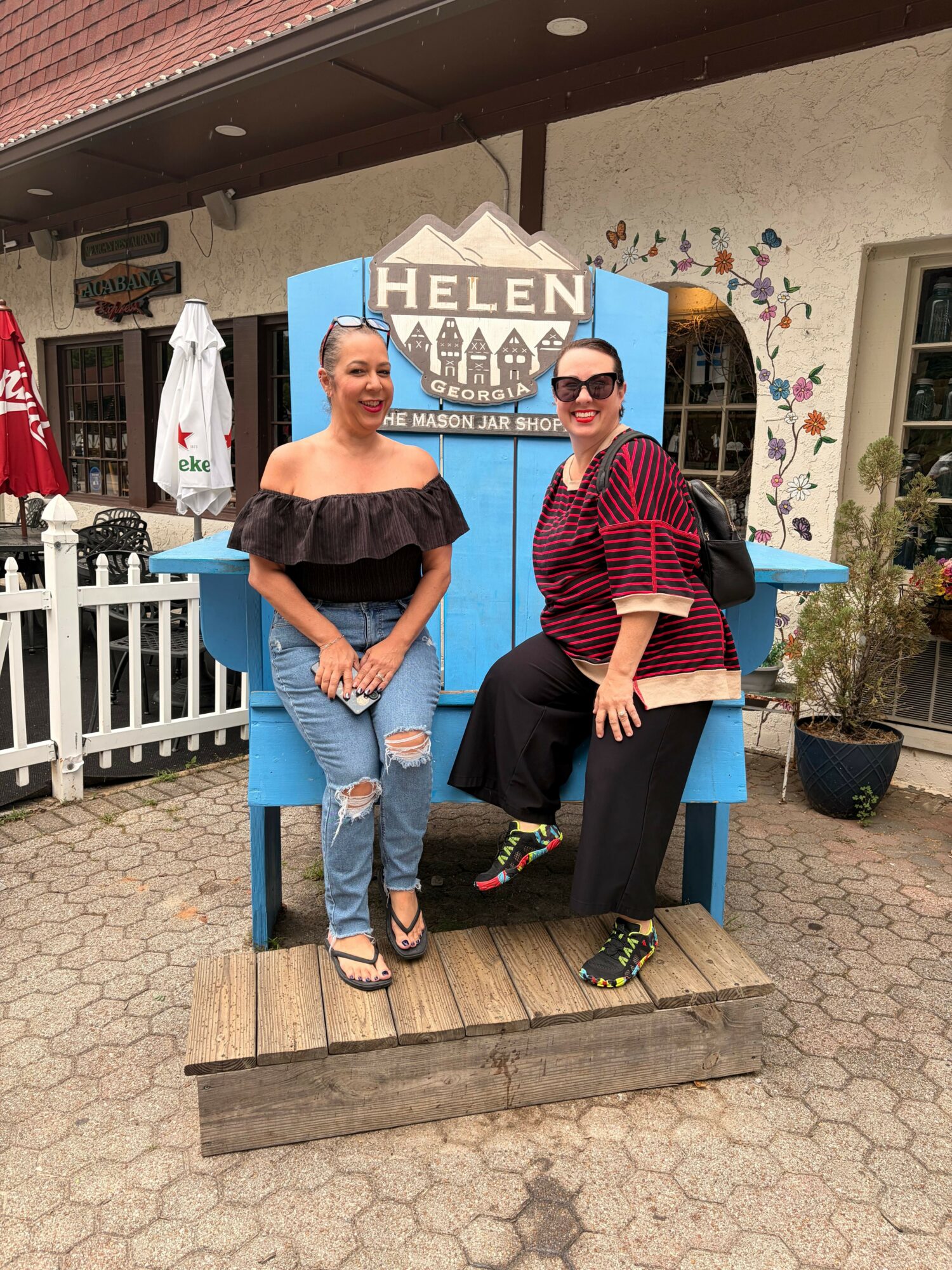 Two women standing in front of a blue photo booth with a sign that says 'HELENA'. One woman wears a striped top and black pants, the other wears a black off-shoulder top and ripped jeans.