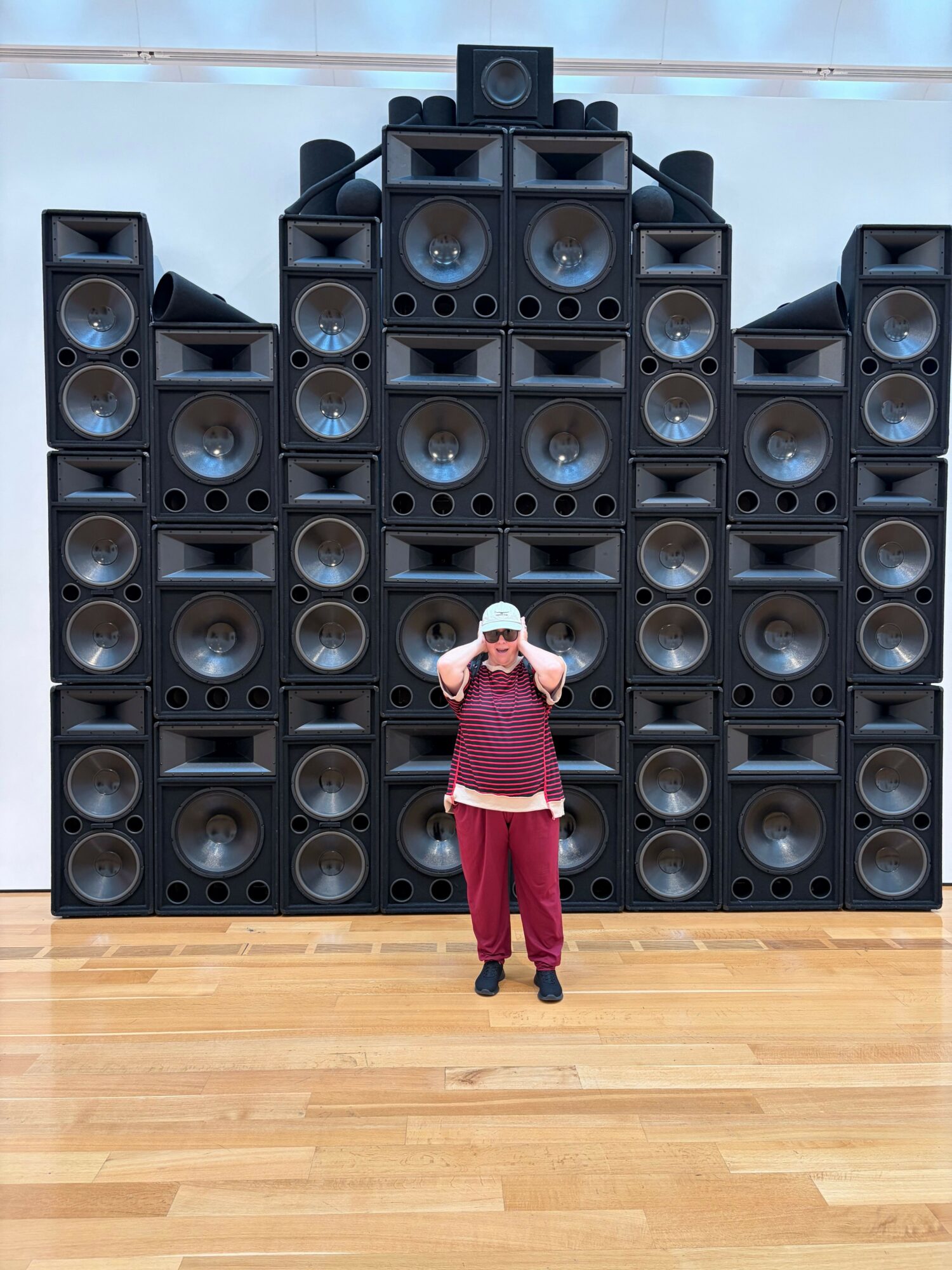 Person in red pants and white shirt standing in front of large speaker system, holding a microphone, on wooden floor.
