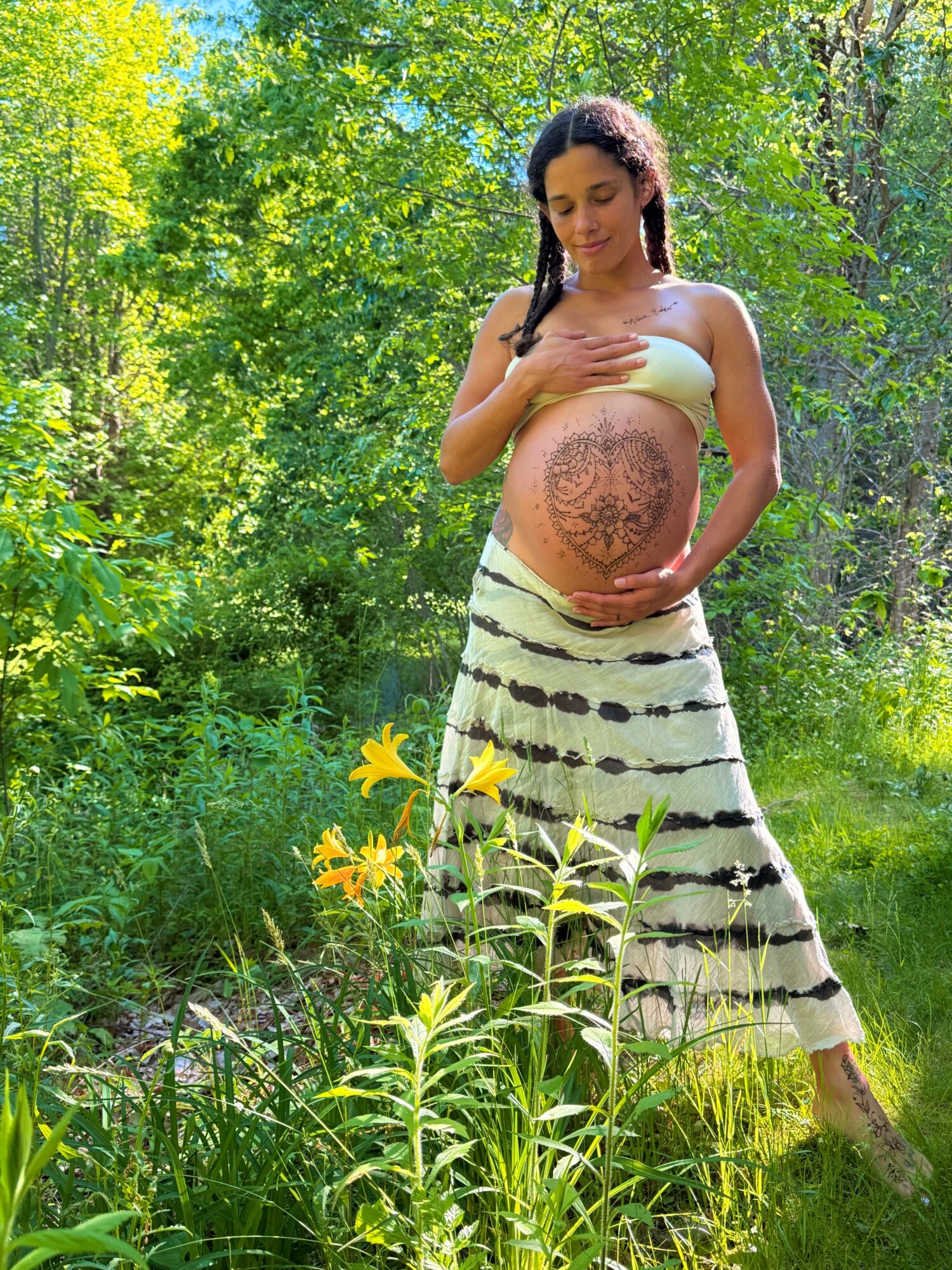 Pregnant woman standing outdoors in a green forest, holding her belly with a detailed tattoo, wearing a striped skirt and a strapless top.