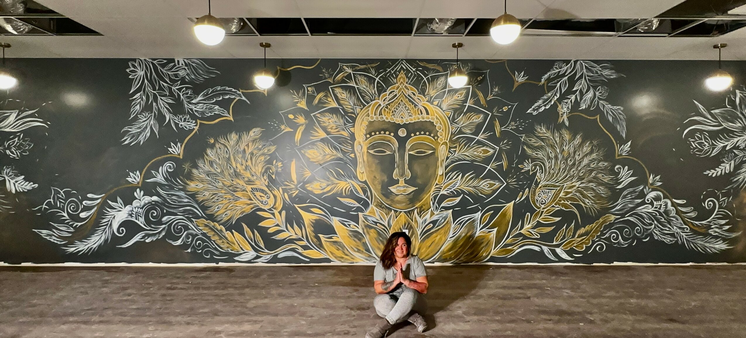 Person sitting in front of a large mural of a golden face with surrounding floral and decorative patterns, ceiling lights overhead.