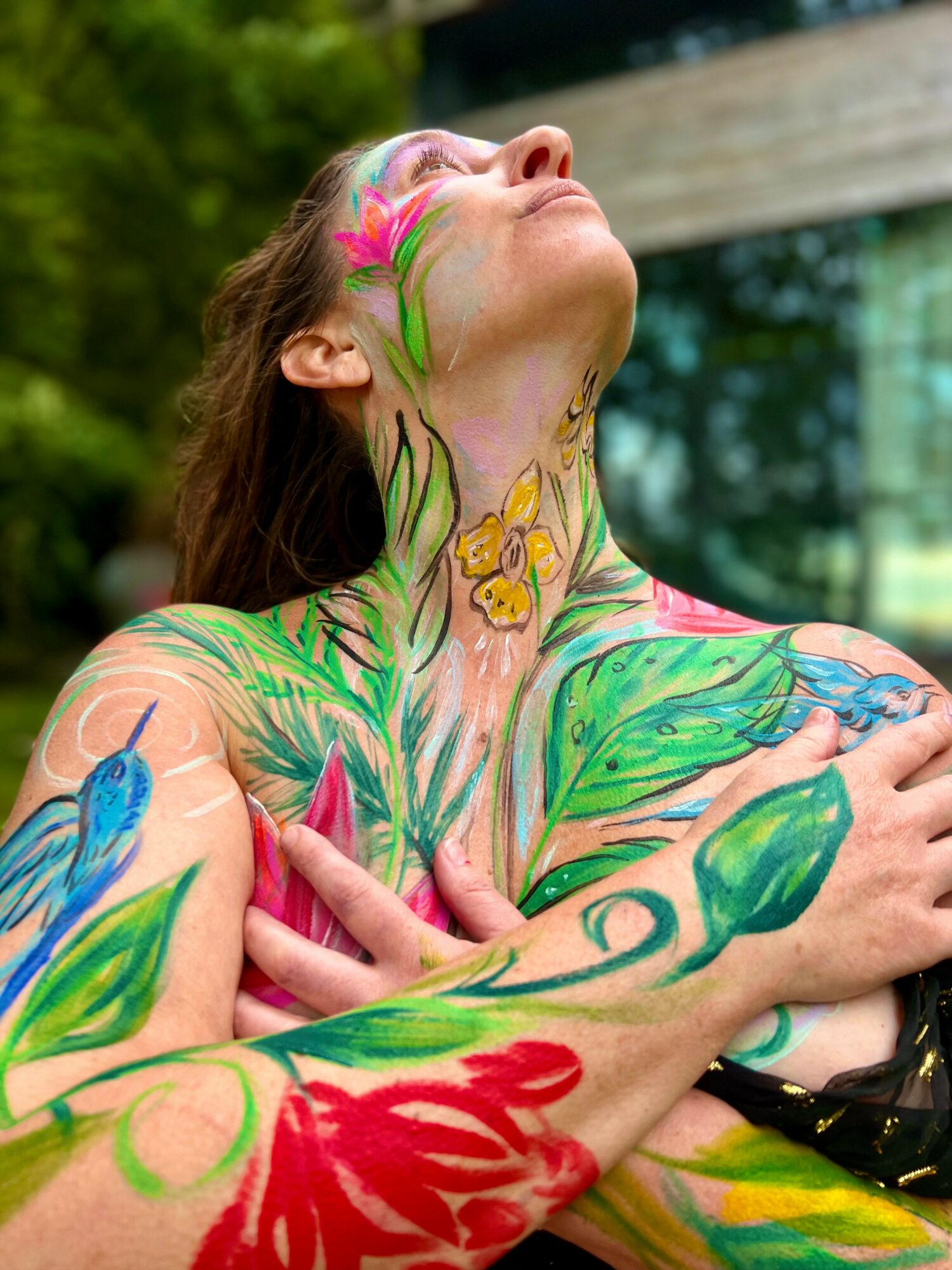 Person with colorful body paint, head tilted back, outdoors, with trees and a building in background.