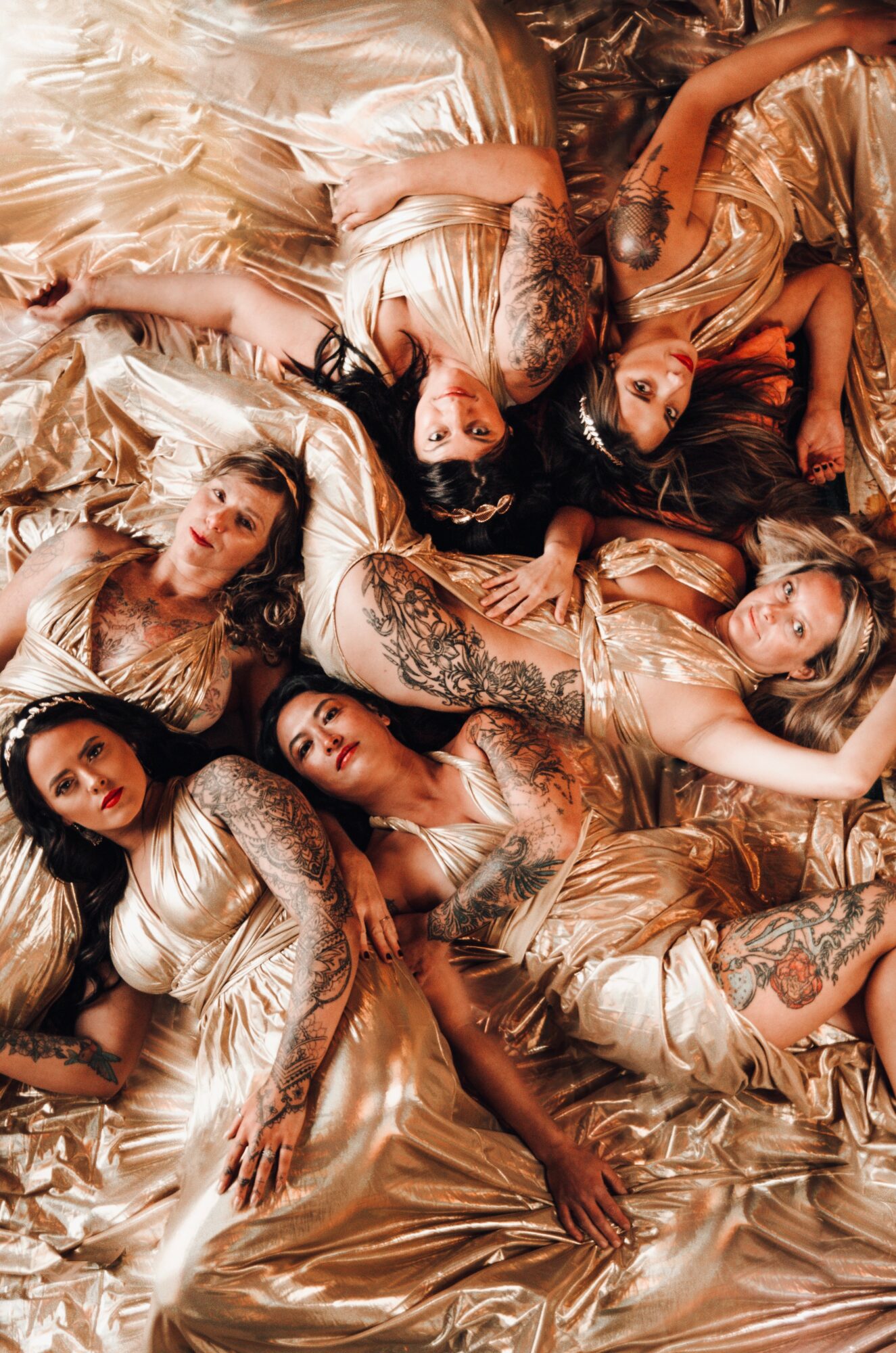 Seven women in gold dresses lying on a shiny surface, arranged in a circular pattern with heads close together.