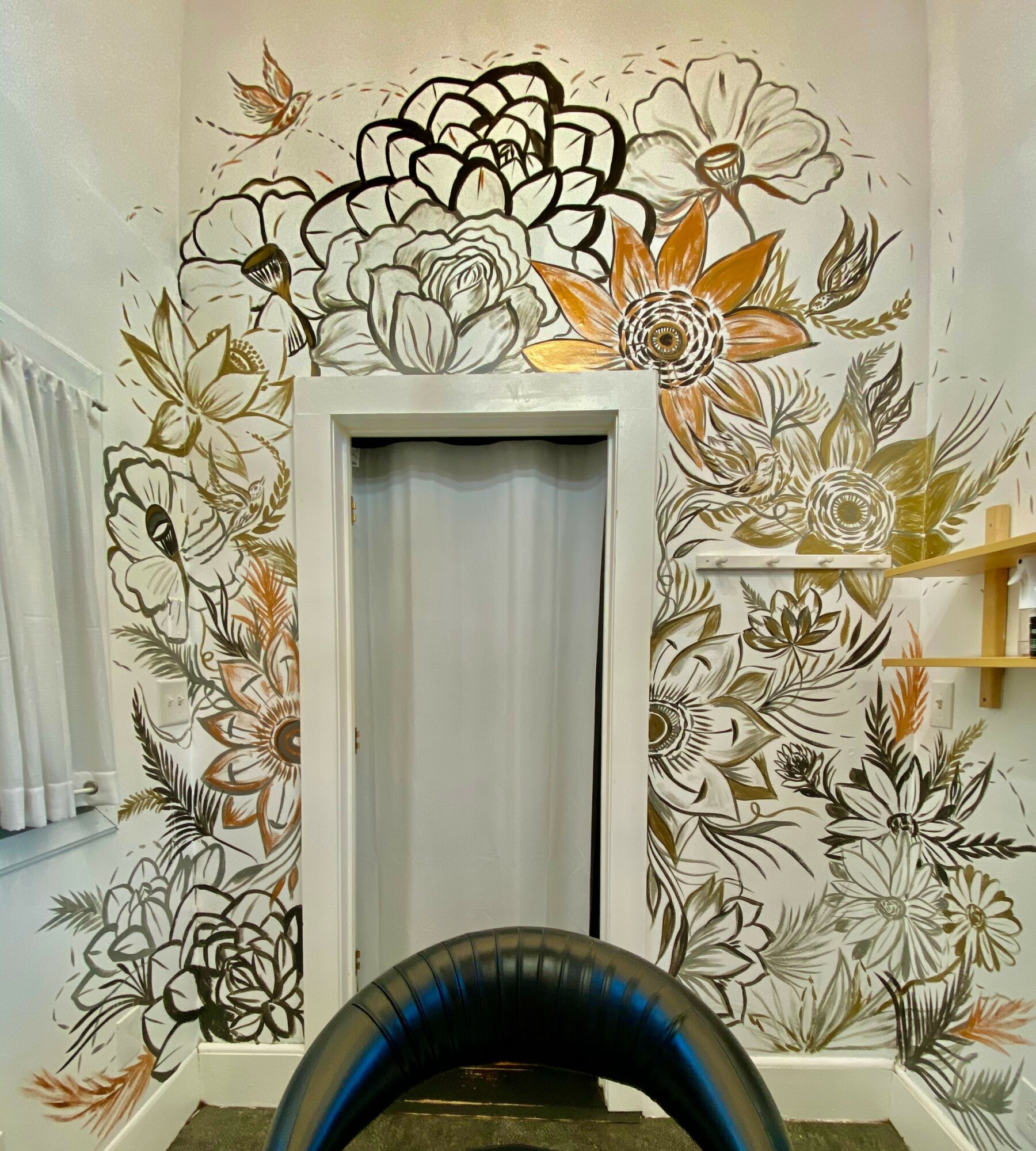 Wall with floral mural surrounding a door, featuring large flowers and leaves in black, white, and gold tones, with shelves on the right.