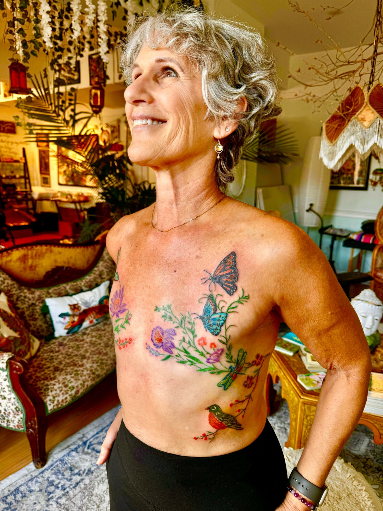 Smiling elderly woman with short gray hair, tattoos of a butterfly, flowers, and a dragon on her chest and side, in a cozy room.