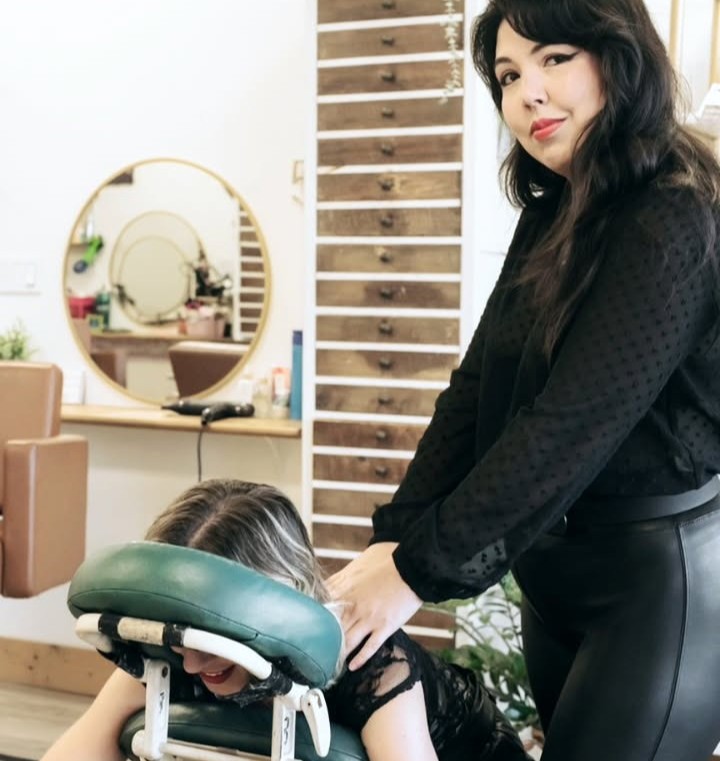 Woman with dark hair and black outfit holding a person's head in a salon, mirror and wooden wall in background.
