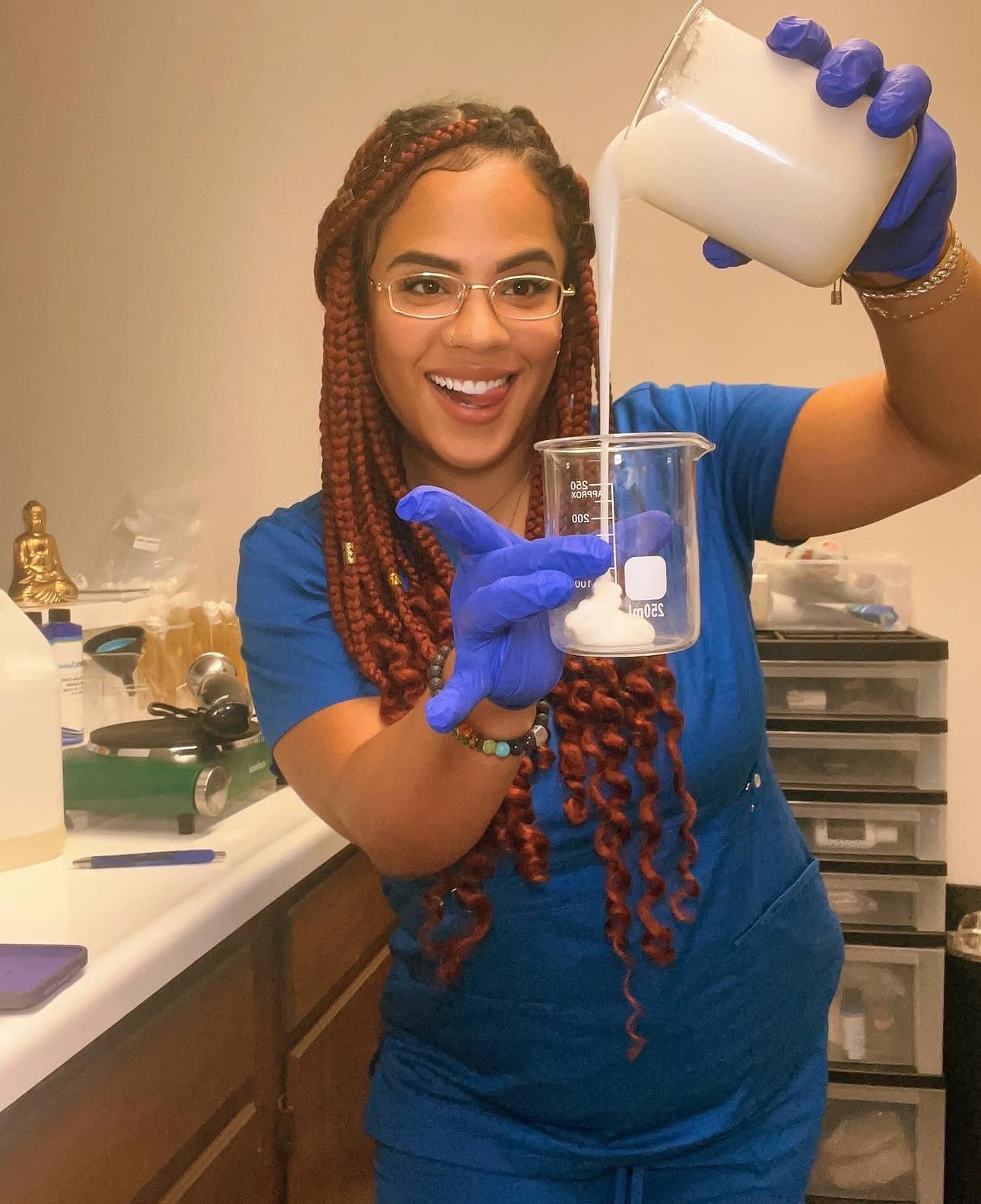 Woman in blue scrubs pouring liquid into a beaker, smiling, wearing glasses and blue gloves, in a laboratory setting.