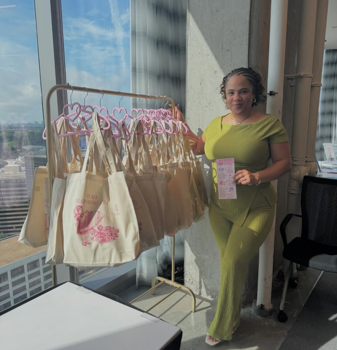 Woman in green outfit standing next to a clothing rack with beige bags, holding a pink ticket, in a high-rise building.