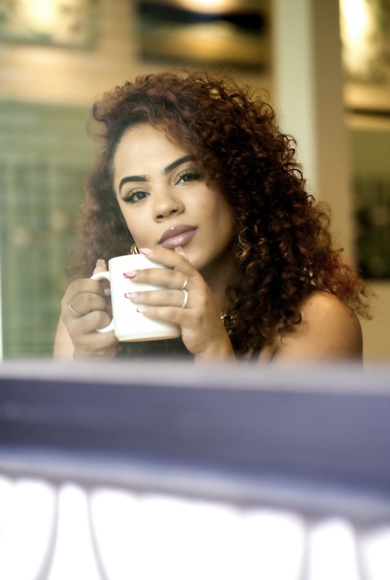 Woman with curly hair holding a white mug, looking at the camera, in a bright indoor setting.