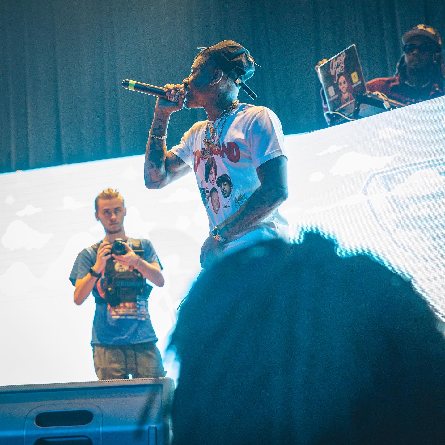 Performer with tattoos and cap singing into microphone on stage, with two people in background, one with a tablet, and a large screen behind.