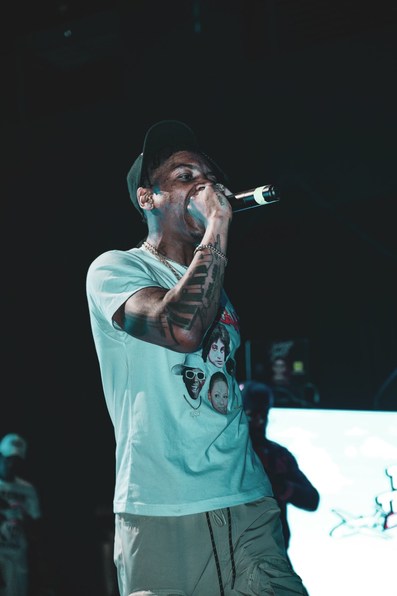 Person with tattoos wearing a cap and graphic t-shirt, holding a microphone, performing on stage in a dark setting.