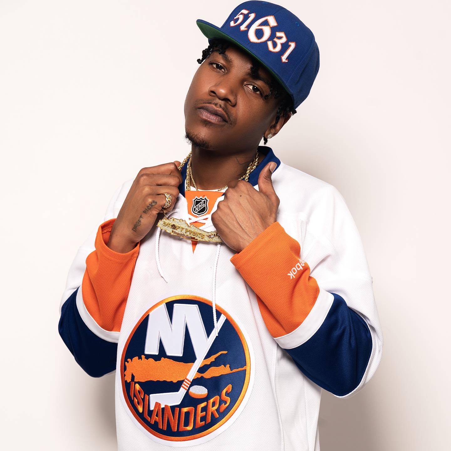 Young man wearing a blue cap, white and blue sports jersey, and gold chains, posing with a serious expression.