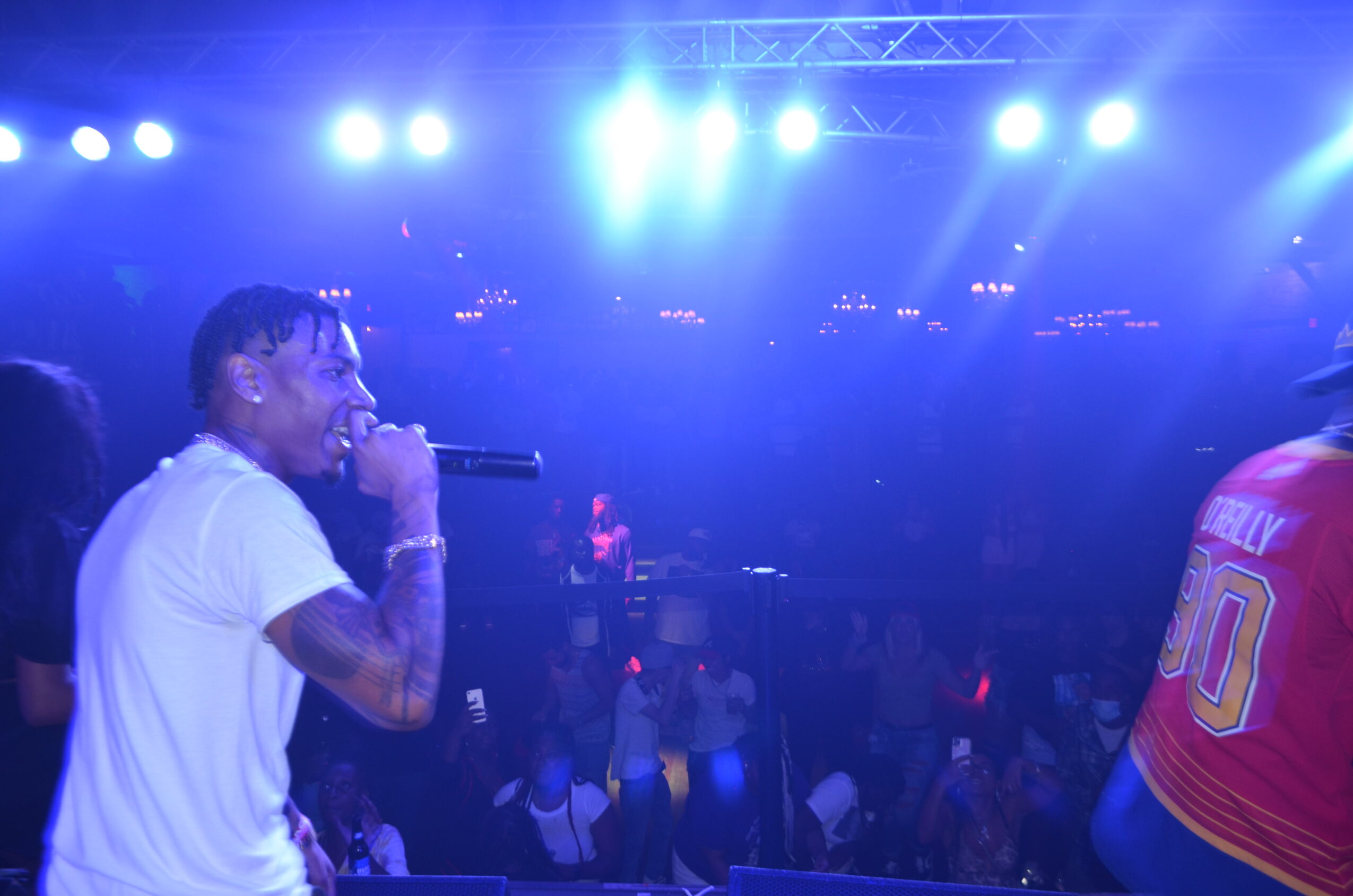 Performer with tattoos singing into a microphone on stage with bright blue lights, audience in the background.
