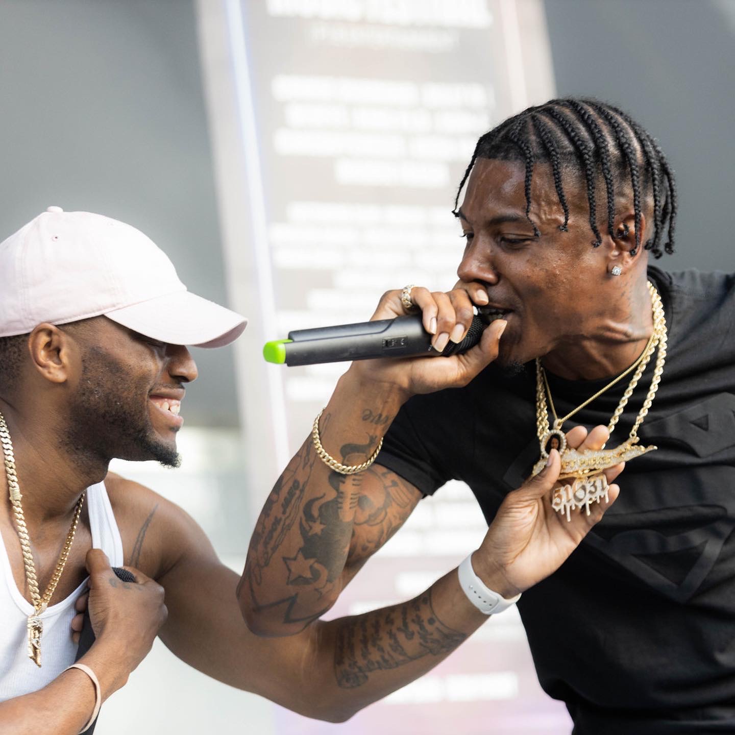 Two men smiling and performing on stage, one holding a microphone, with a blurred background. Word tattoo visible on arm.