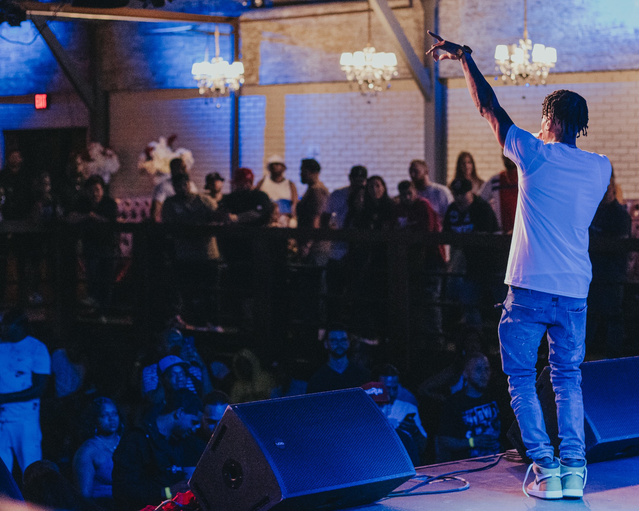 Performer on stage with arm raised, audience watching in a dimly lit venue with chandeliers.