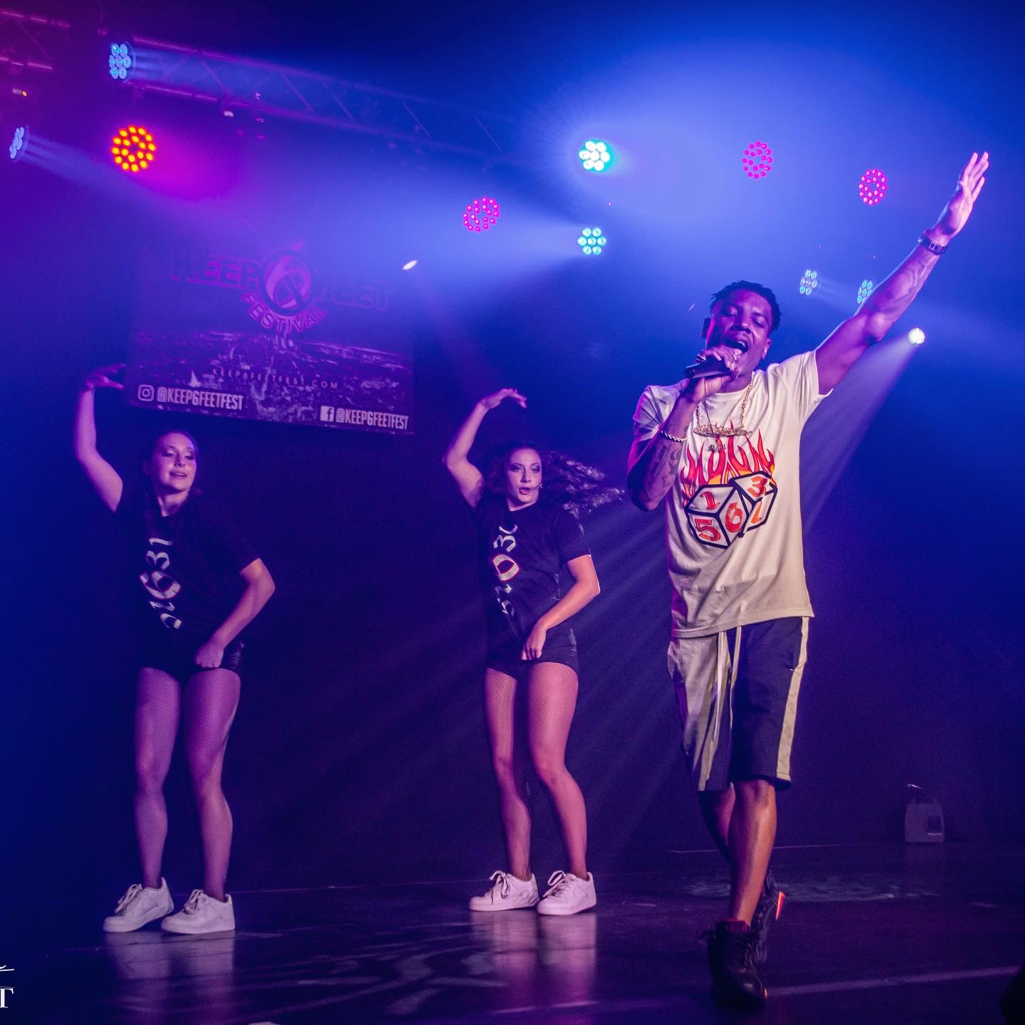 Performer singing into microphone with two dancers behind on stage, colorful lights overhead, dark background.