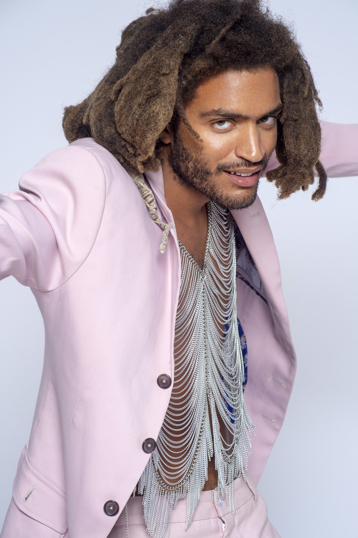 Man with dreadlocks wearing a pink blazer and layered necklaces, smiling at the camera.
