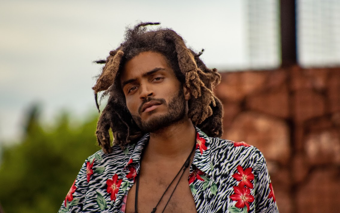 Young man with dreadlocks and a patterned shirt outdoors, with a blurred background of trees and buildings.