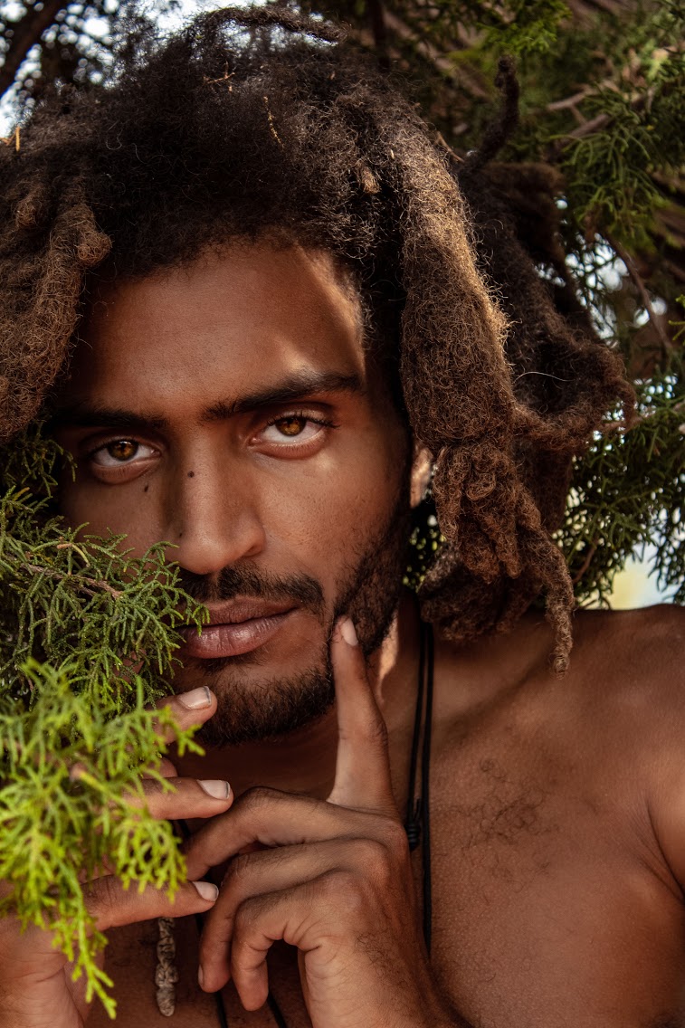 Close-up of a person with dreadlocks, intense gaze, and hand near face, outdoors with greenery in background.