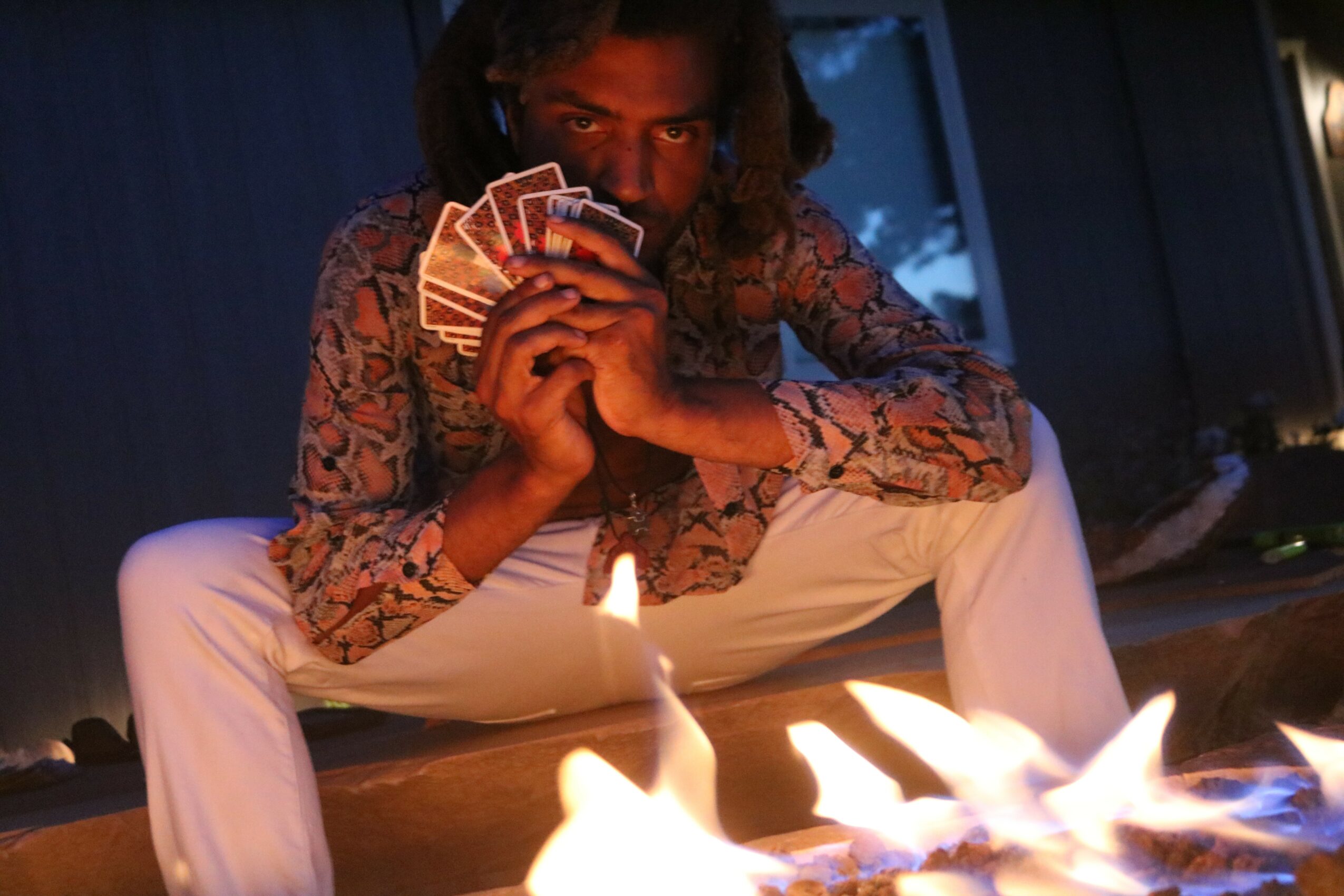 Person sitting outdoors at night holding a fan of playing cards, with flames in front of them.