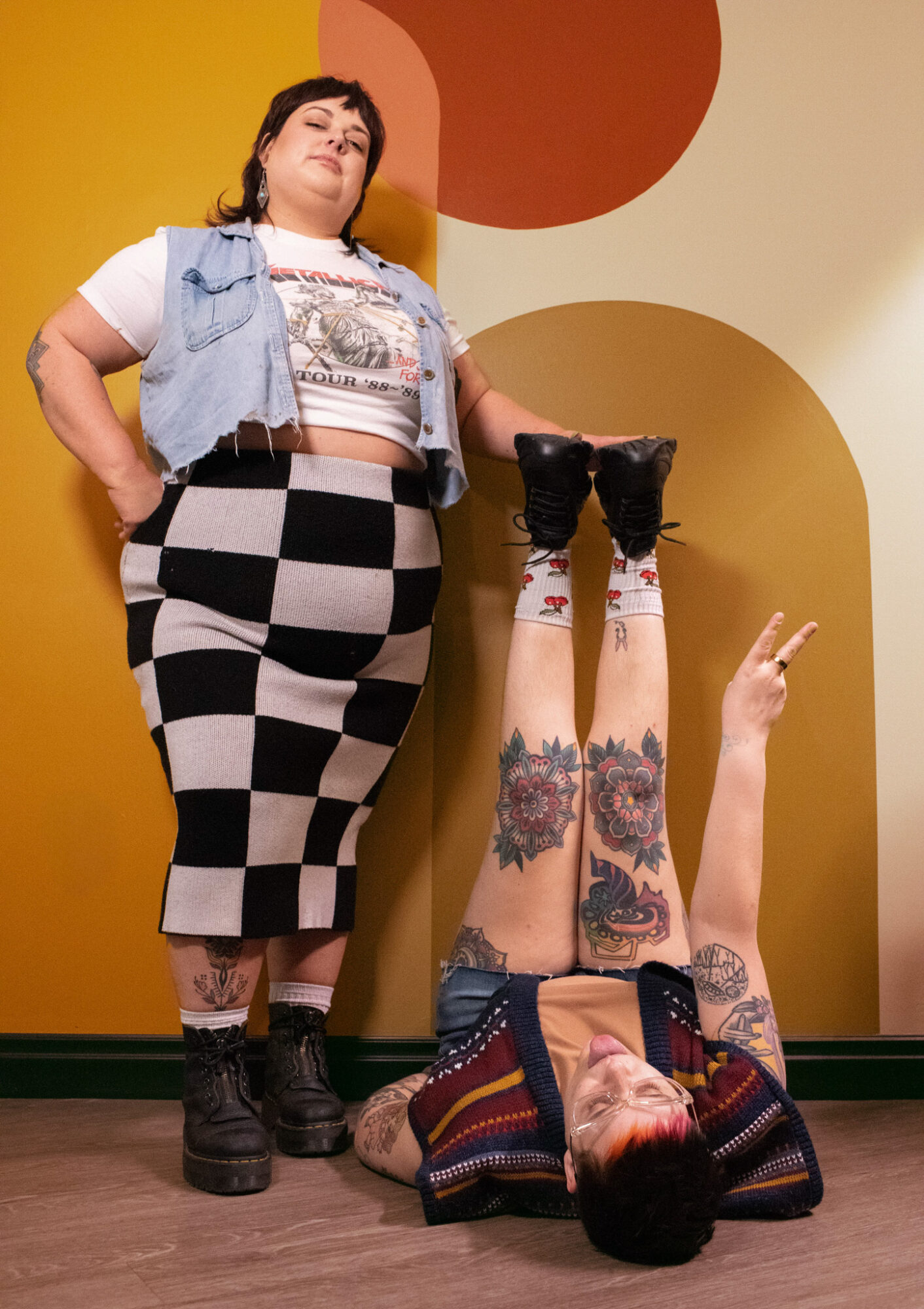 Two women pose against a colorful wall; one stands with hands on hips, the other lies on the floor making a peace sign.