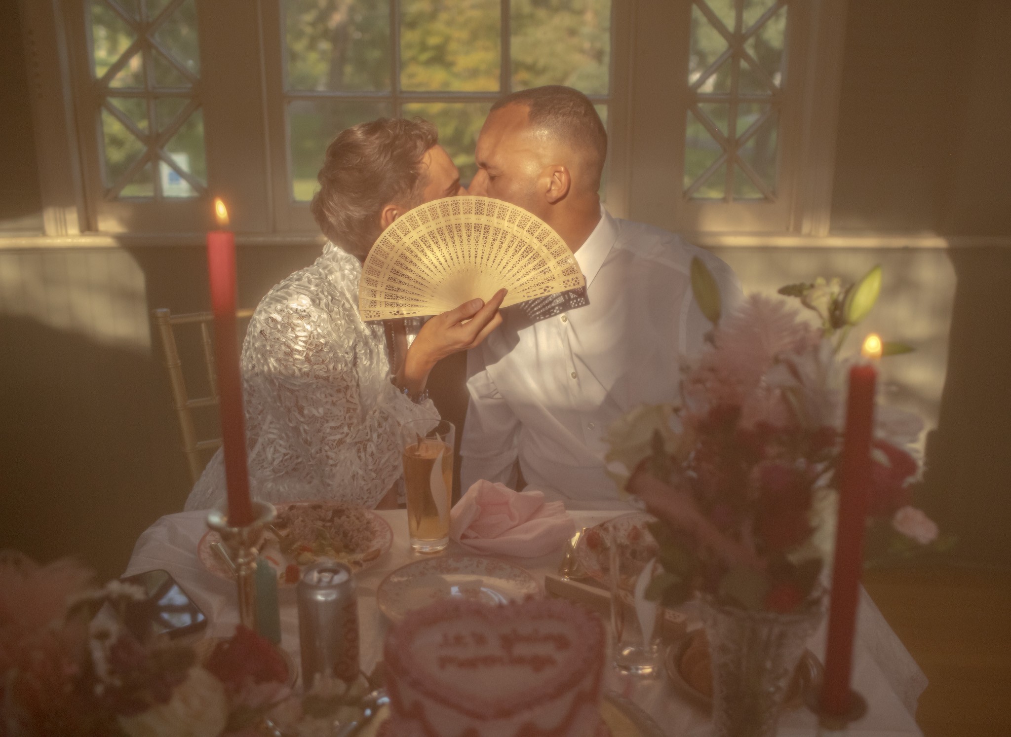 Two people sit at a table with candles, flowers, and a cake, sharing a kiss behind a fan held by one person.