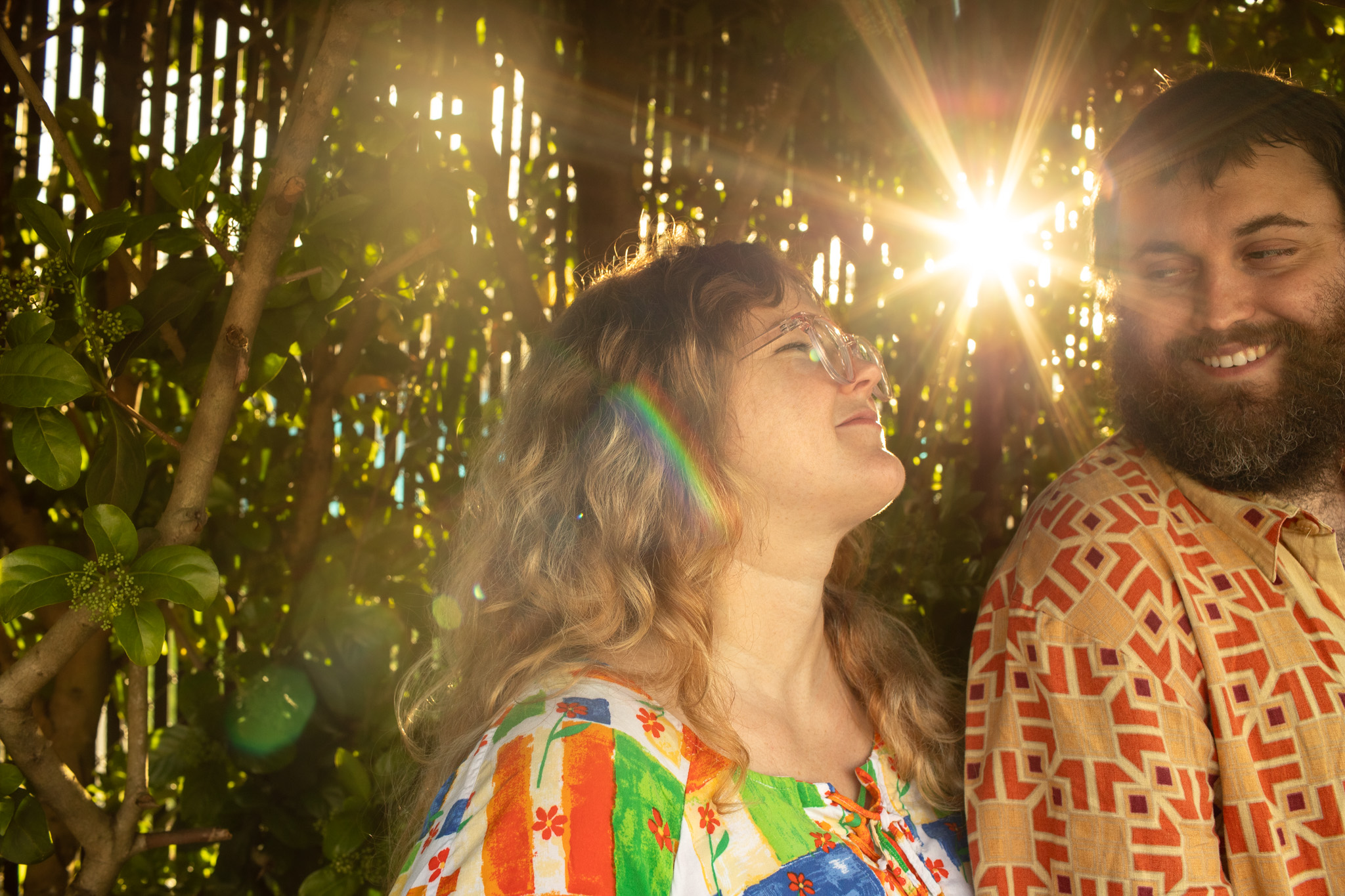 Two people smiling outdoors with sunlight shining through trees behind them.