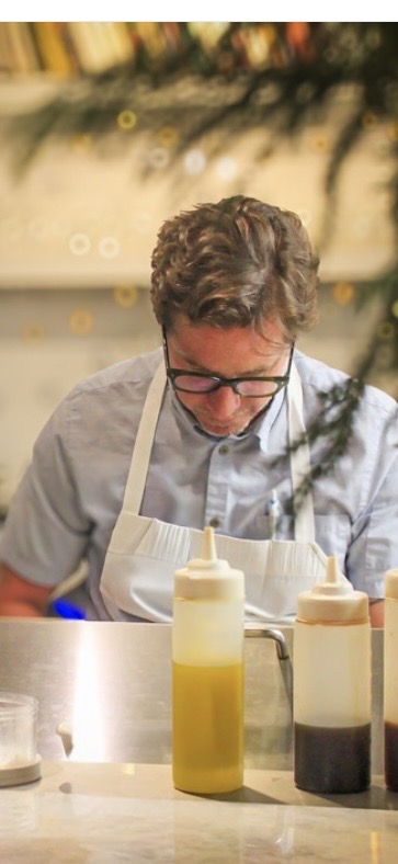 Man with glasses and curly hair wearing an apron, working with bottles on a table.