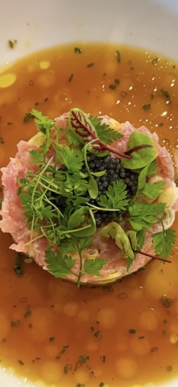 A dish with a pink, textured base topped with fresh green herbs and microgreens, served in a bowl with orange sauce.