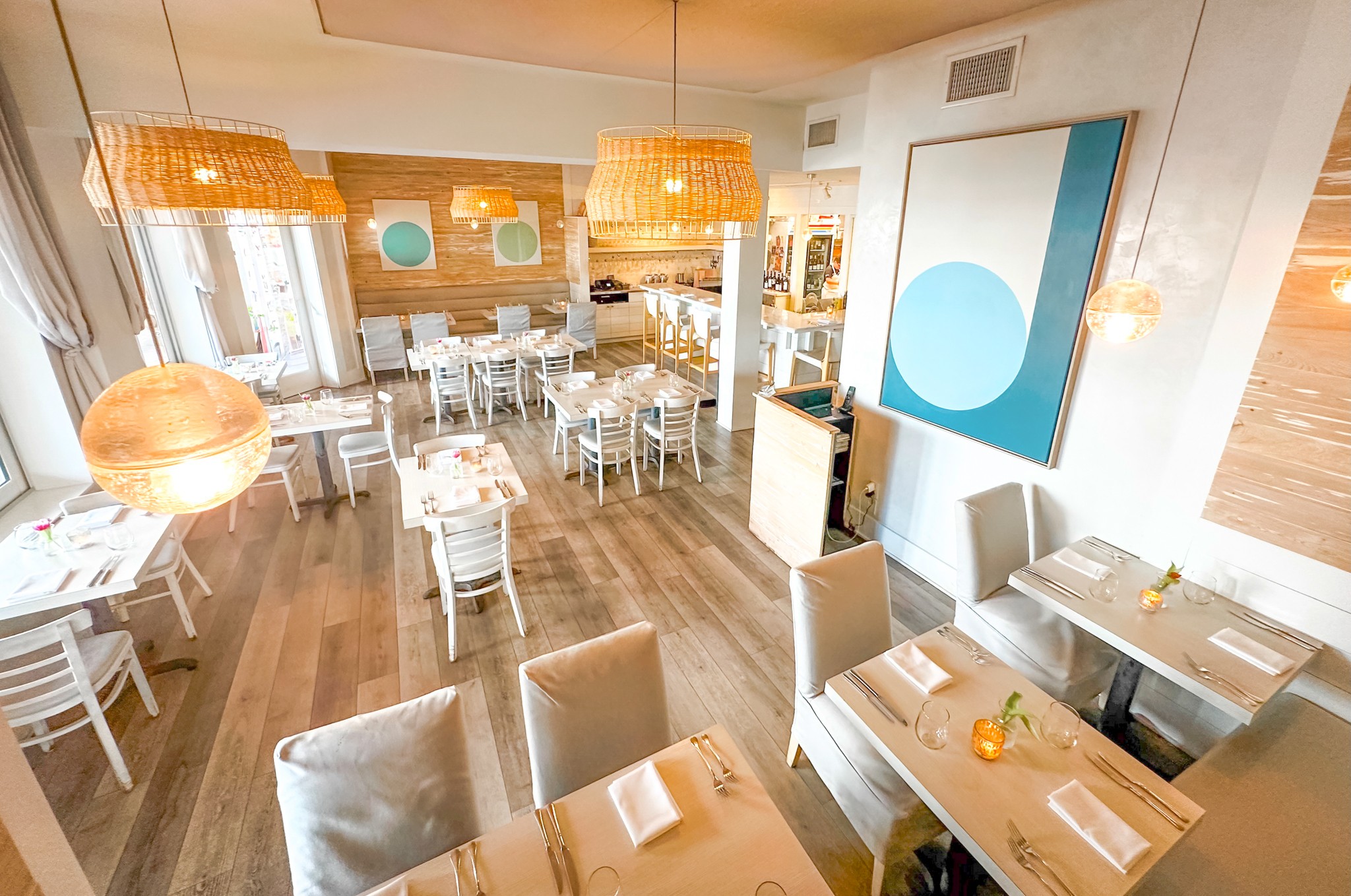 Bright restaurant interior with wooden floors, white chairs, large hanging lamps, and modern artwork on the wall.