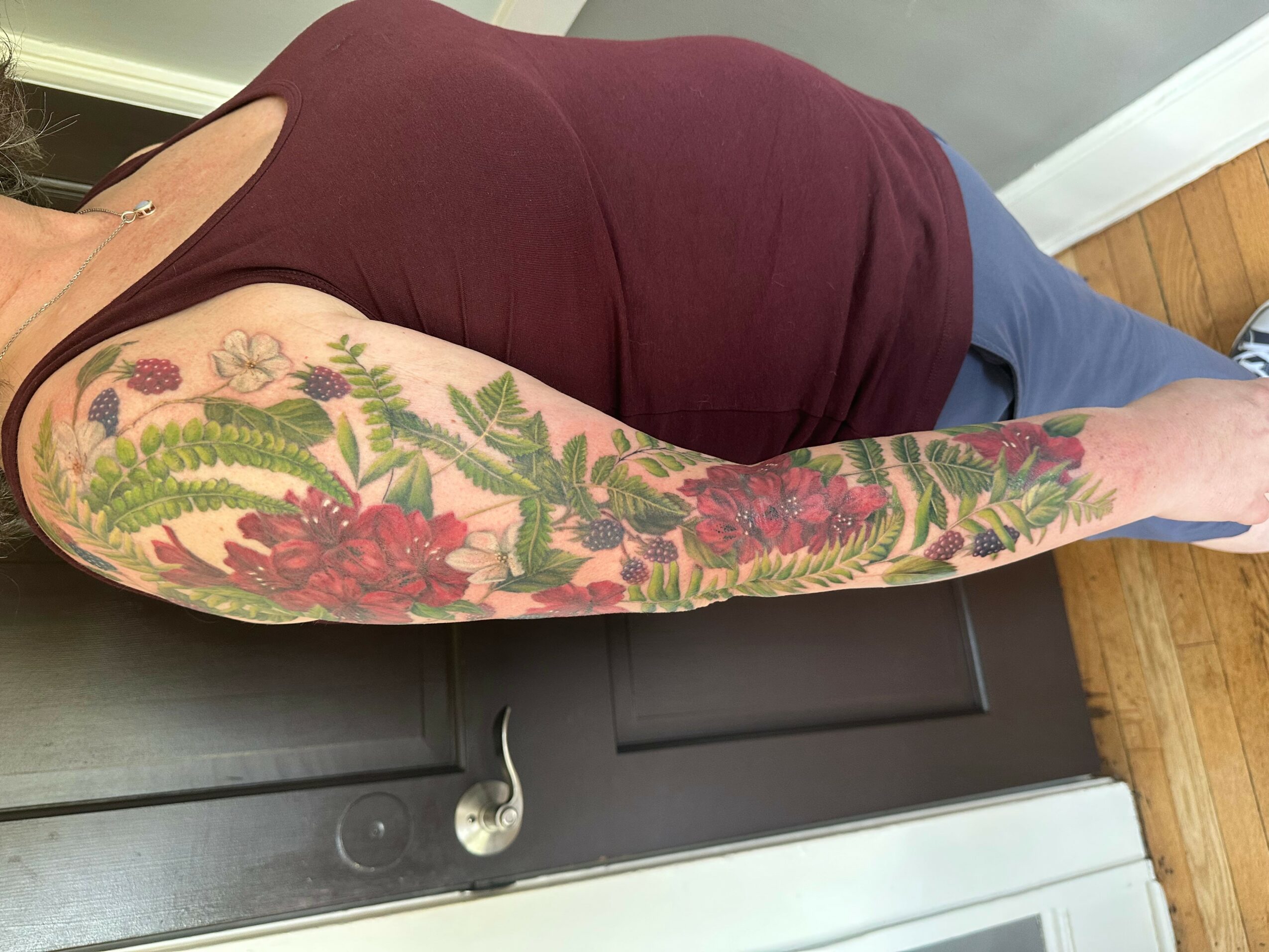 Side view of a person with a floral tattoo on their arm, wearing a maroon top and blue pants, standing indoors.