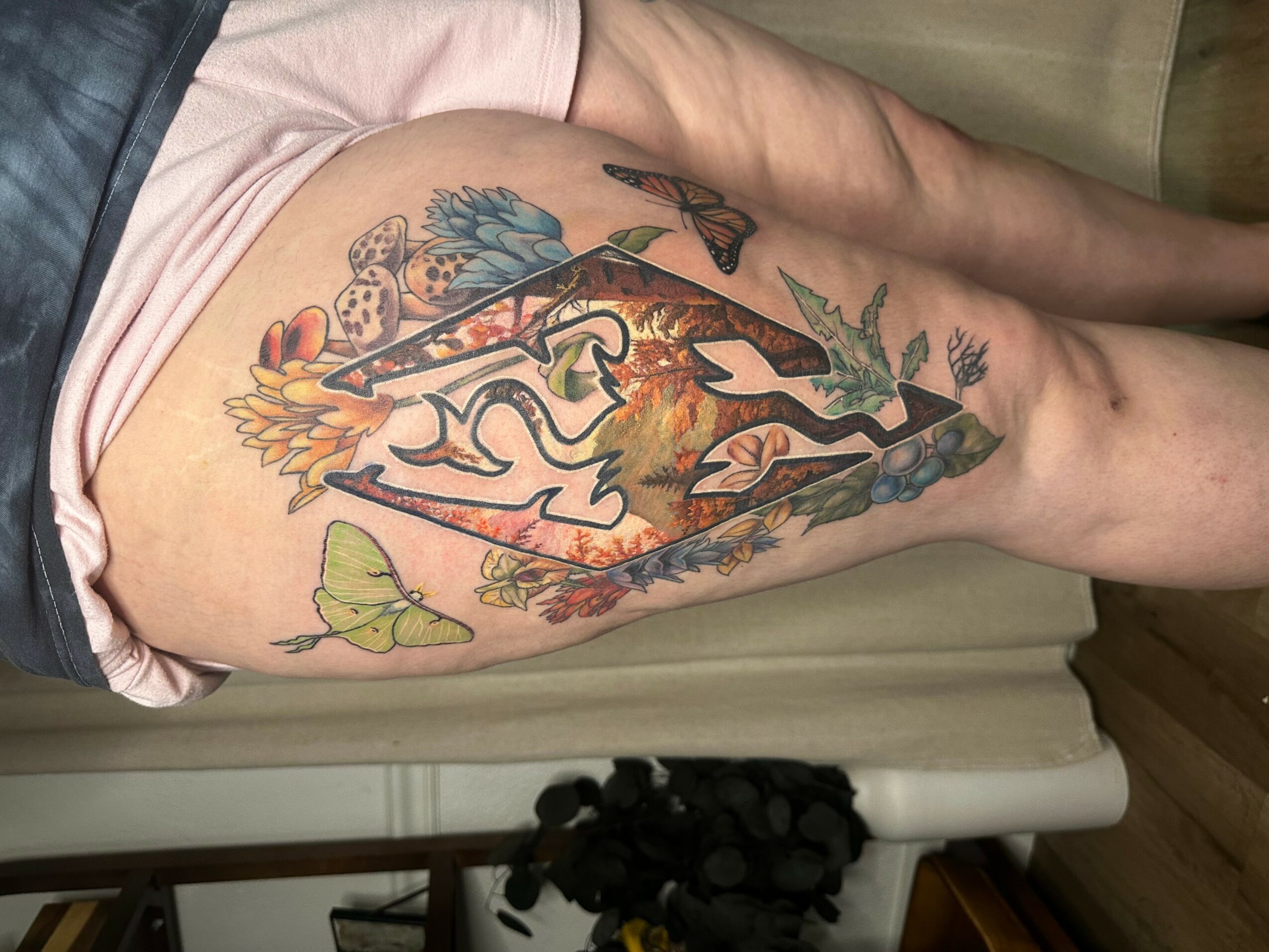 Tattoo of a fox with flowers, leaves, and butterflies on person's thigh, with a background of trees and nature elements.