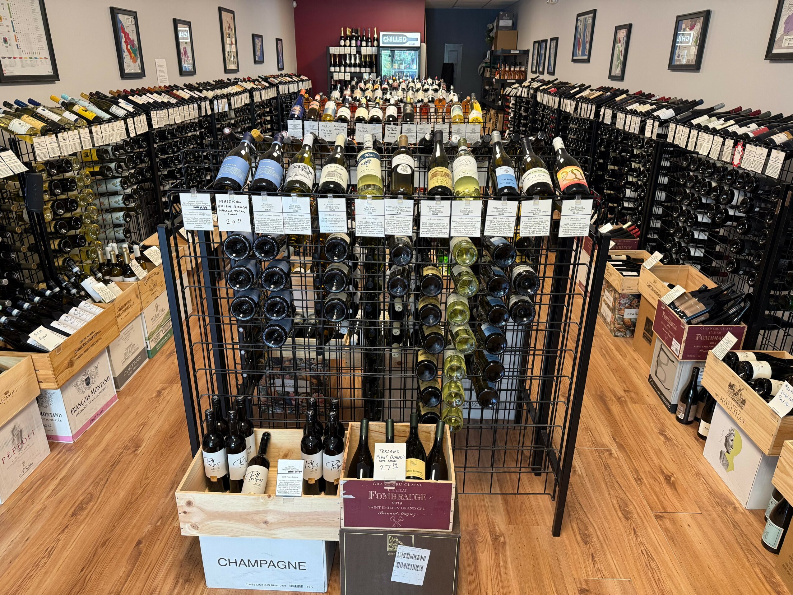 Wine bottles displayed on racks in a wine shop with shelves of bottles on walls, wooden floor, and a customer area in the background.
