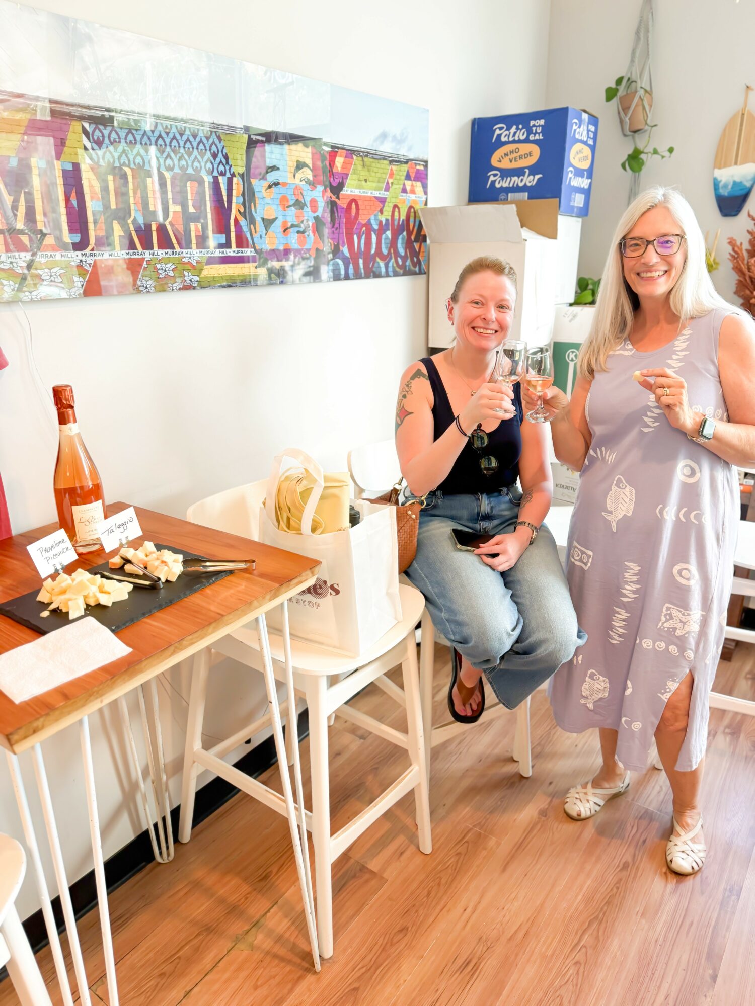 Two women smiling and holding drinks in a decorated room with a table, snacks, and colorful wall art.