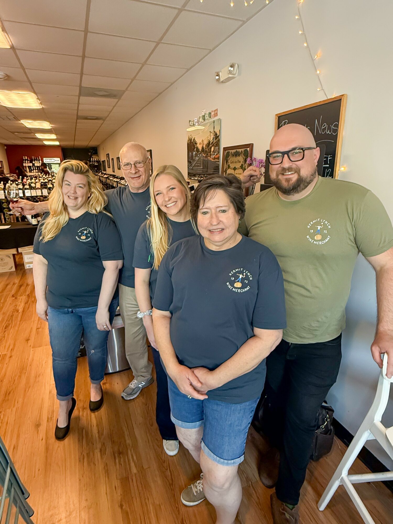 Five people standing in a line indoors, smiling, with a chalkboard and wall decorations behind them.