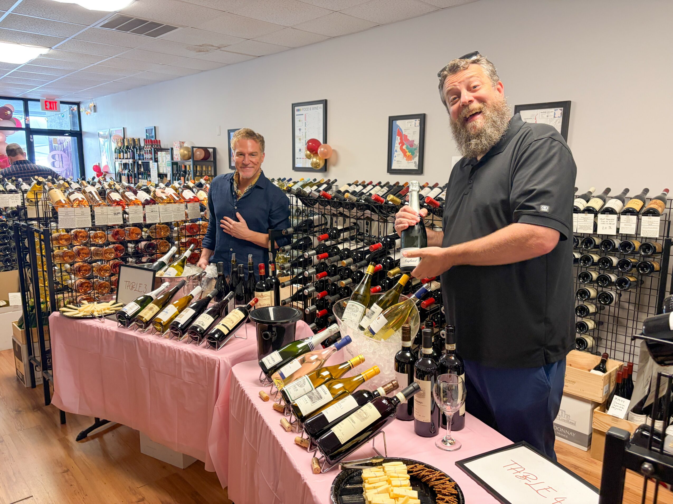 Two men standing behind a wine display table with bottles, in a store with wine racks and artwork on the wall.