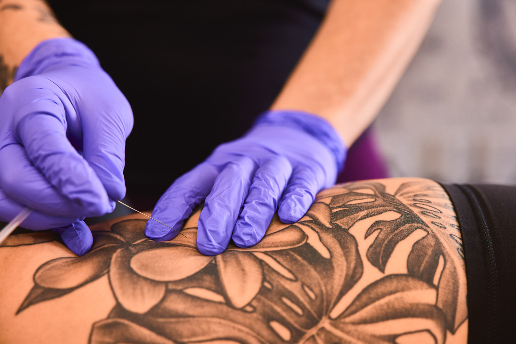 Person with purple gloves tattooing a large floral design on a person's leg.