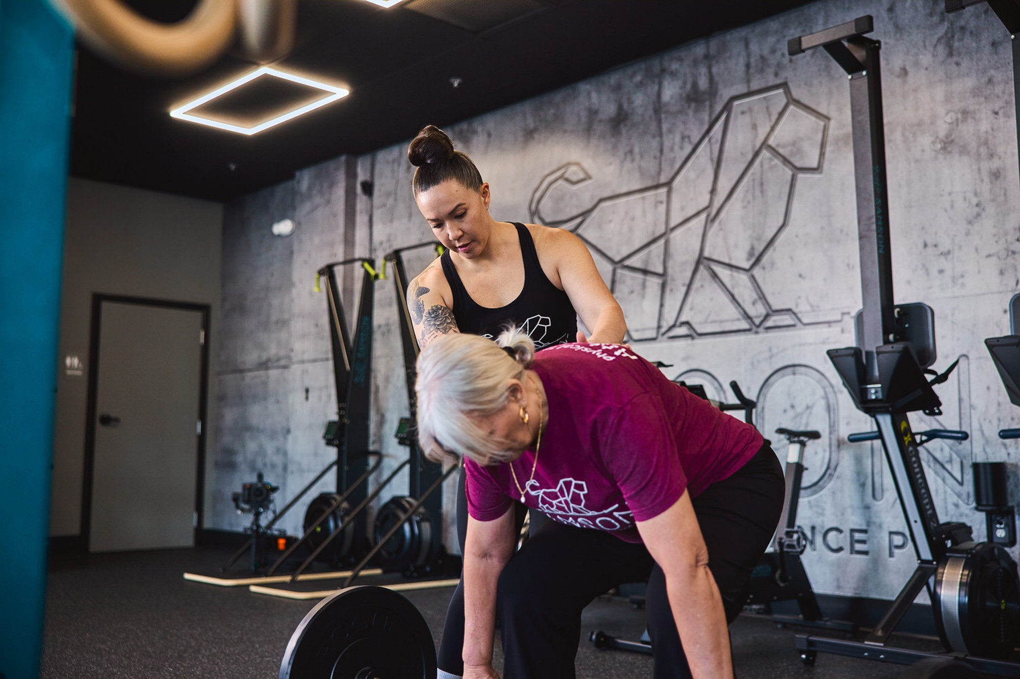 A woman with a bun assists an older woman with gray hair in a gym, both focused on exercise equipment.
