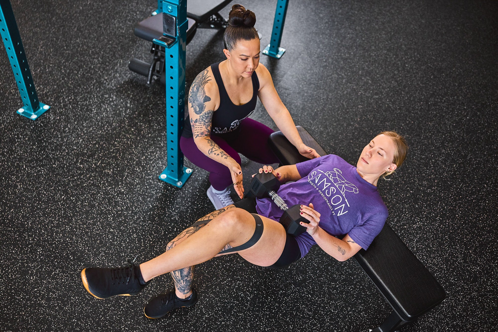 A woman in purple workout clothes lies on a bench while another woman in black and purple assists her with a leg exercise.