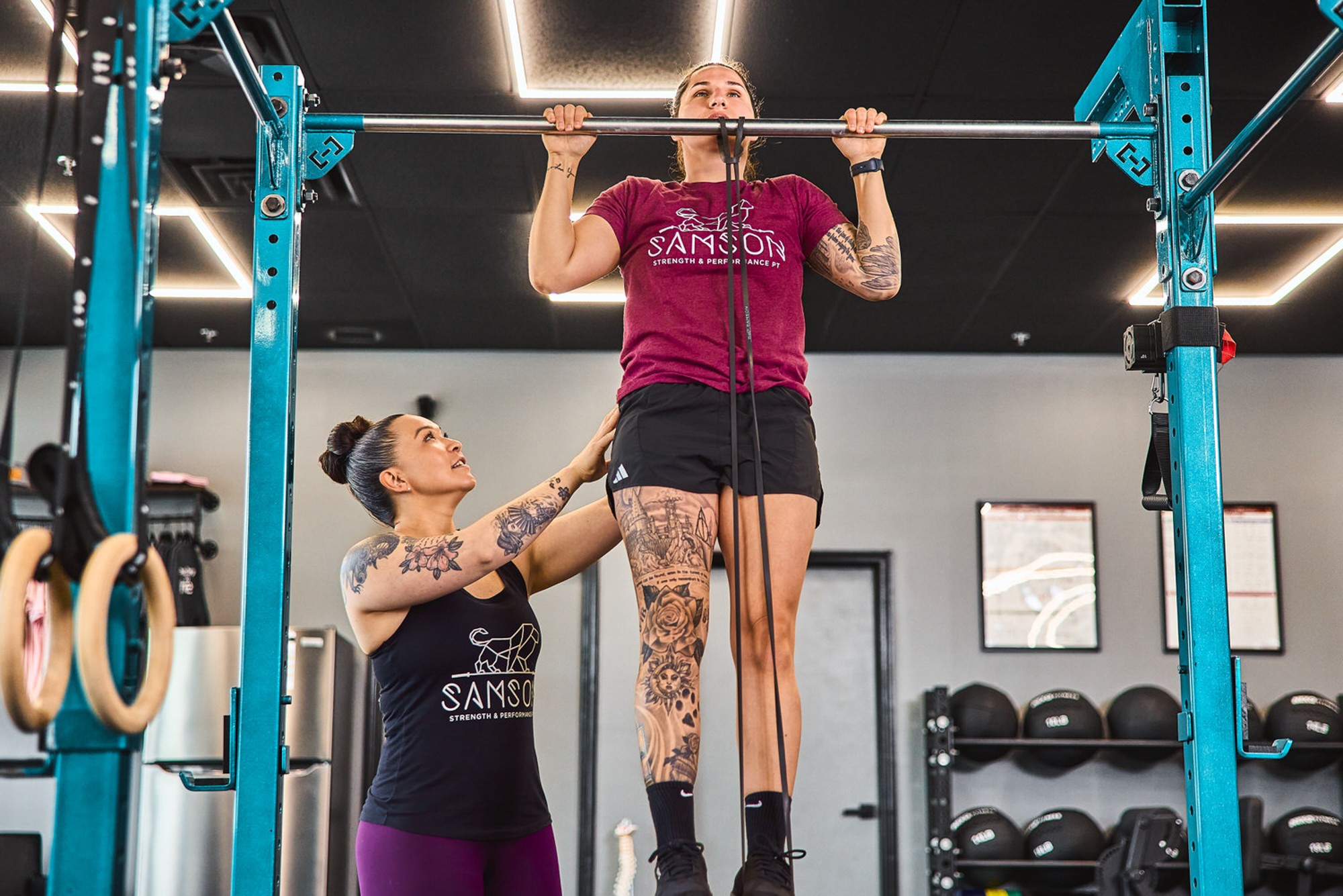 Woman lifting a barbell with assistance from trainer in a gym, black and blue equipment visible, woman wearing a maroon shirt and patterned leggings.