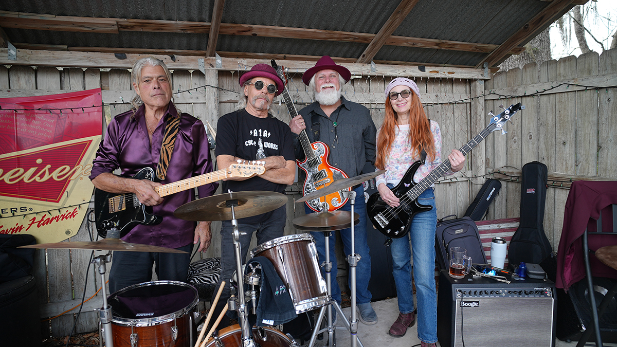 Four musicians with instruments in a backyard, wooden fence background, casual clothing, some wearing hats and sunglasses.