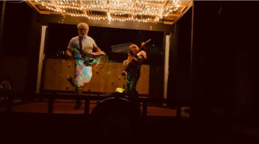 Two musicians perform on stage with a large screen behind them, illuminated by warm lighting.
