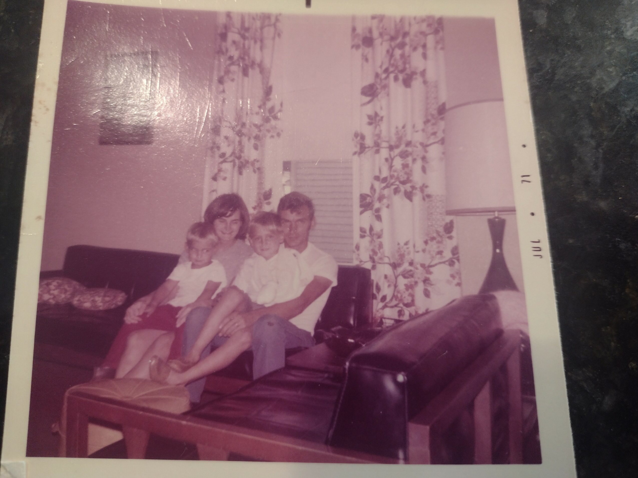 Two children and an adult sitting on a sofa in a living room with curtains and a lamp.