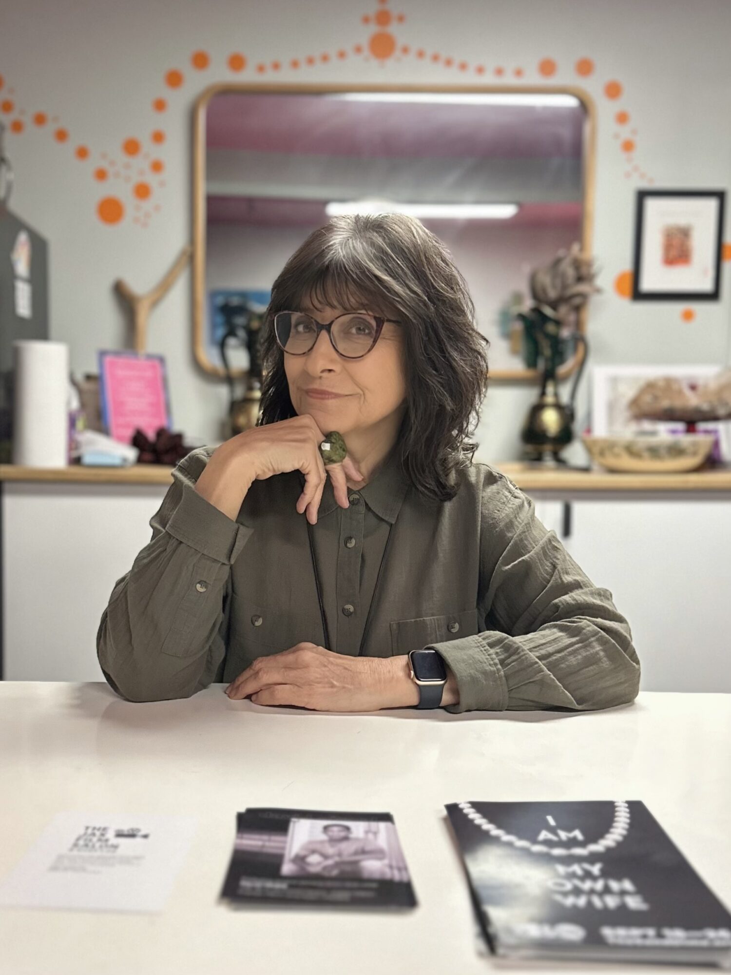 Woman with glasses and wavy hair sitting at a table, resting her chin on her hand, in a decorated room.