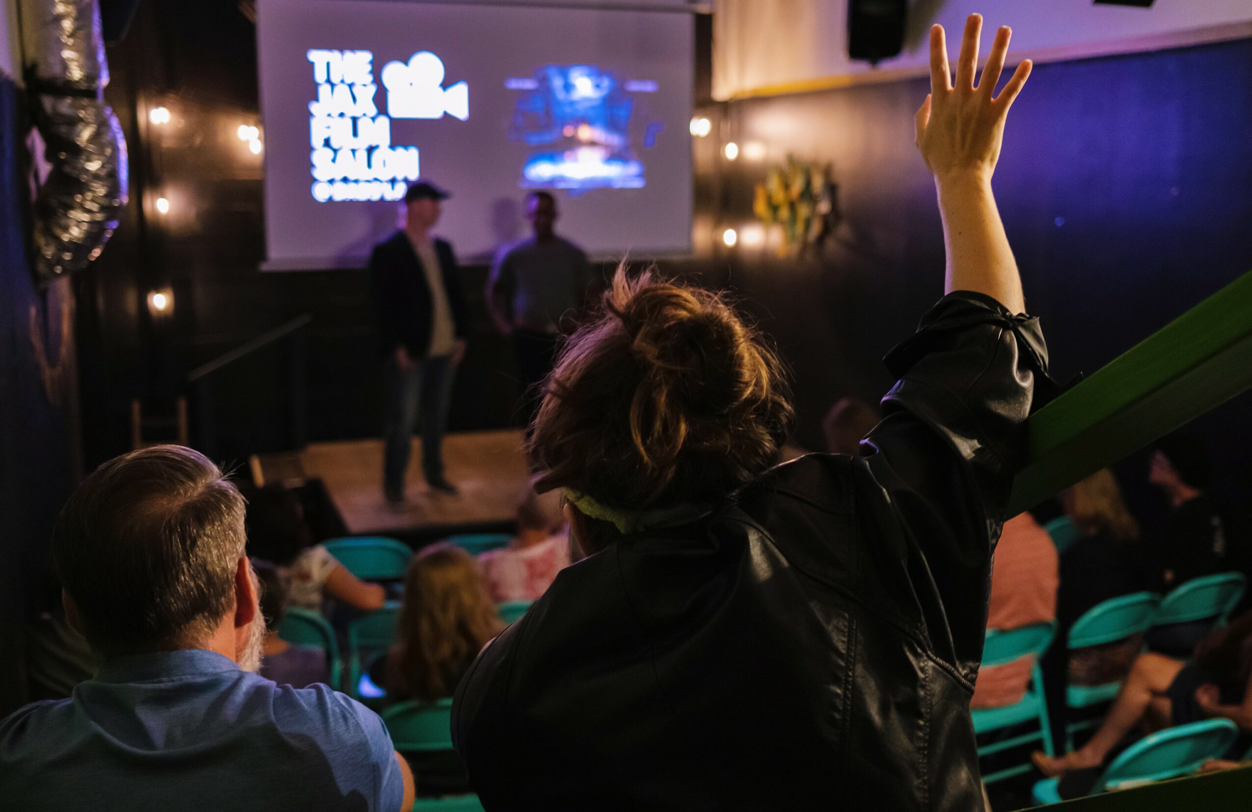 Person raising hand in audience during a presentation or event, with others seated and a screen in the background.